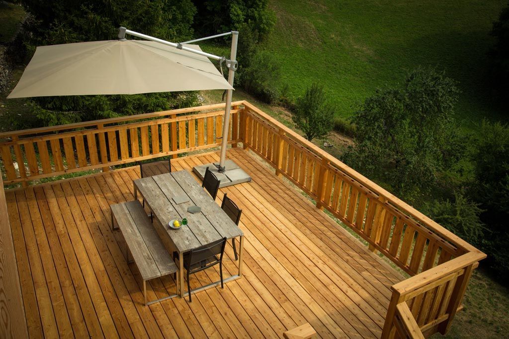 Terrasse en bois avec table, bancs et parasol déporté donnant sur une colline herbeuse.