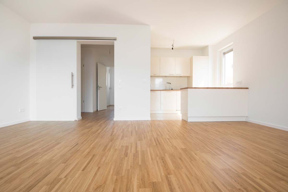Appartement vide avec parquet, murs blancs et un aperçu d'un coin cuisine.