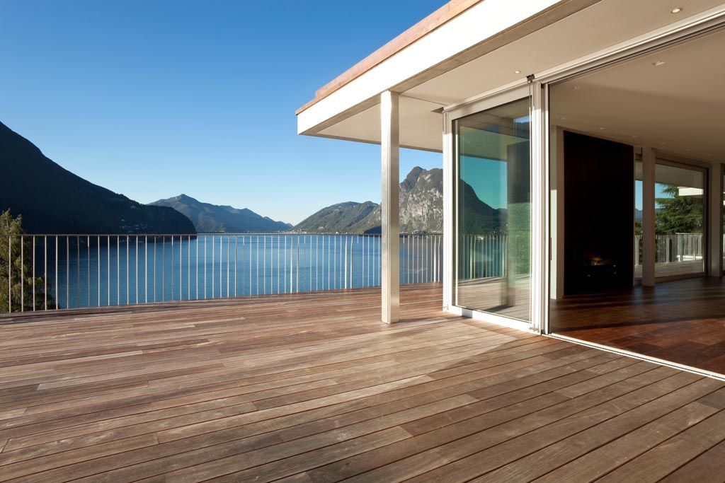 Terrasse en bois d'une maison moderne donnant sur un lac et des montagnes, avec portes coulissantes en verre.