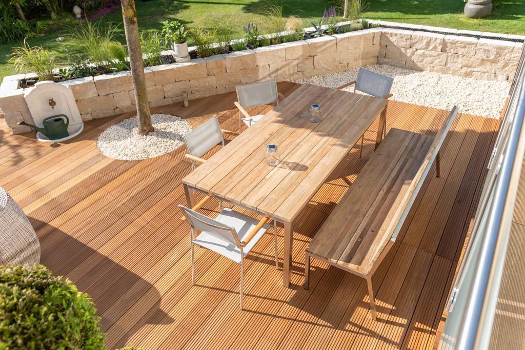 Terrasse en bois avec table, banc et chaises, entourée d'un mur en pierre et d'aménagements paysagers.
