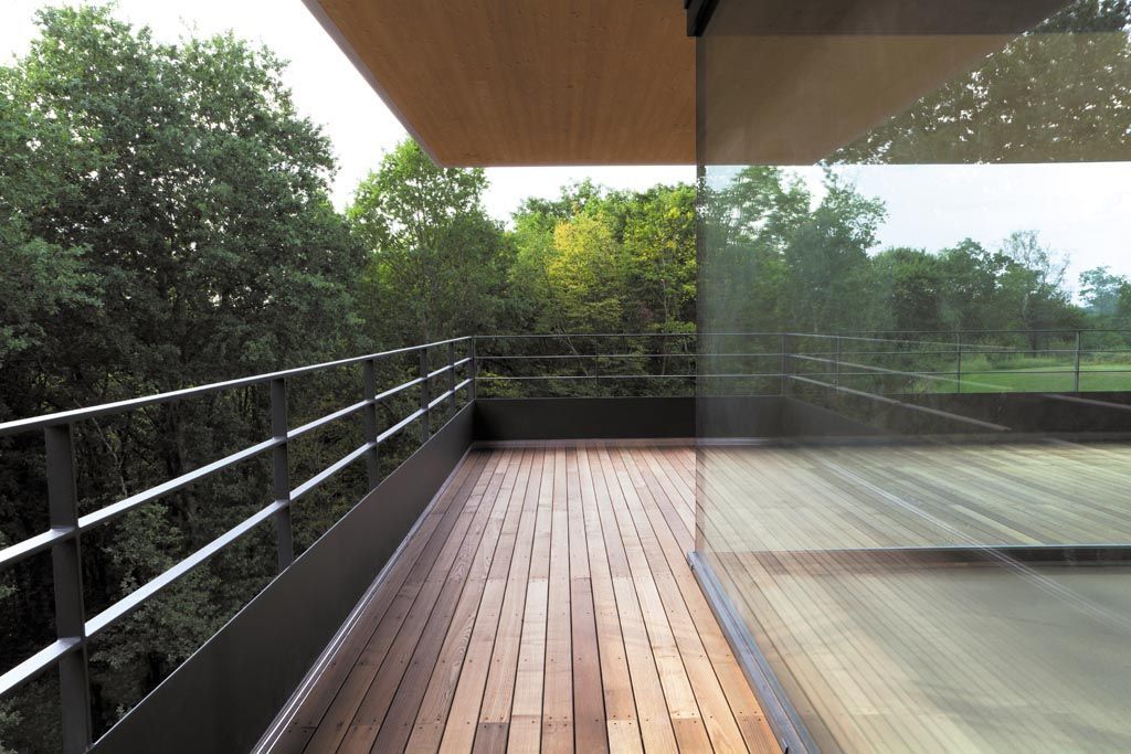 Terrasse en bois avec rambarde en métal, baies vitrées du sol au plafond et vue sur la forêt, reflétant les arbres.