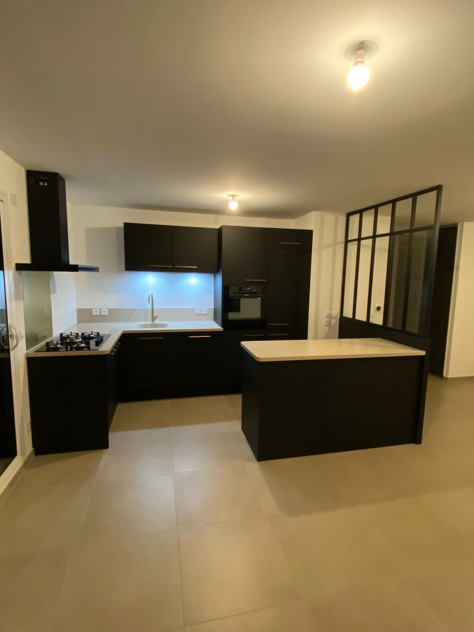 Cuisine moderne noire avec îlot central, plan de travail blanc, électroménager en inox et grandes baies vitrées.