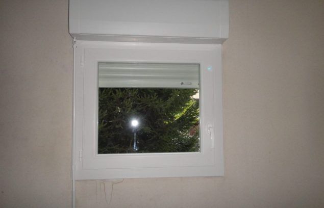 Fenêtre à cadre blanc avec store fermé, laissant apparaître un arbre et une lumière. Fixée sur un mur beige.