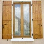 Fenêtre à volets en bois ouverts, encastrée dans un mur clair. Rideaux intérieurs visibles à travers la vitre.