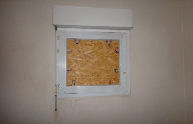 Une fenêtre condamnée par des planches, encadrée de blanc, sur un mur beige, avec un auvent blanc.