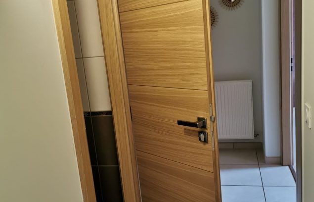 Une porte en bois entrouverte laisse entrevoir une salle de bains avec un radiateur et un sol carrelé.