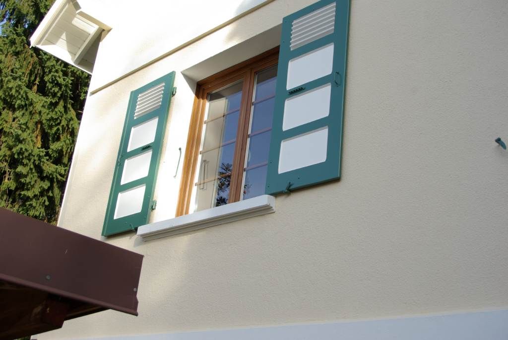 Maison couleur crème avec une fenêtre encadrée de bois, des volets verts et des touches de blanc.