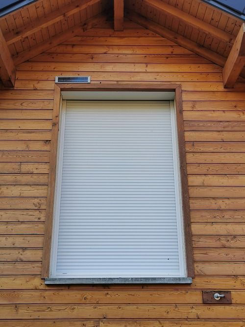 Fenêtre avec volet roulant blanc fermé sur une maison en bois.