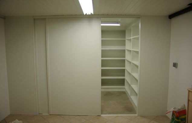 Une porte coulissante blanche dévoile un garde-manger avec des étagères blanches et un luminaire, sur un sol beige.