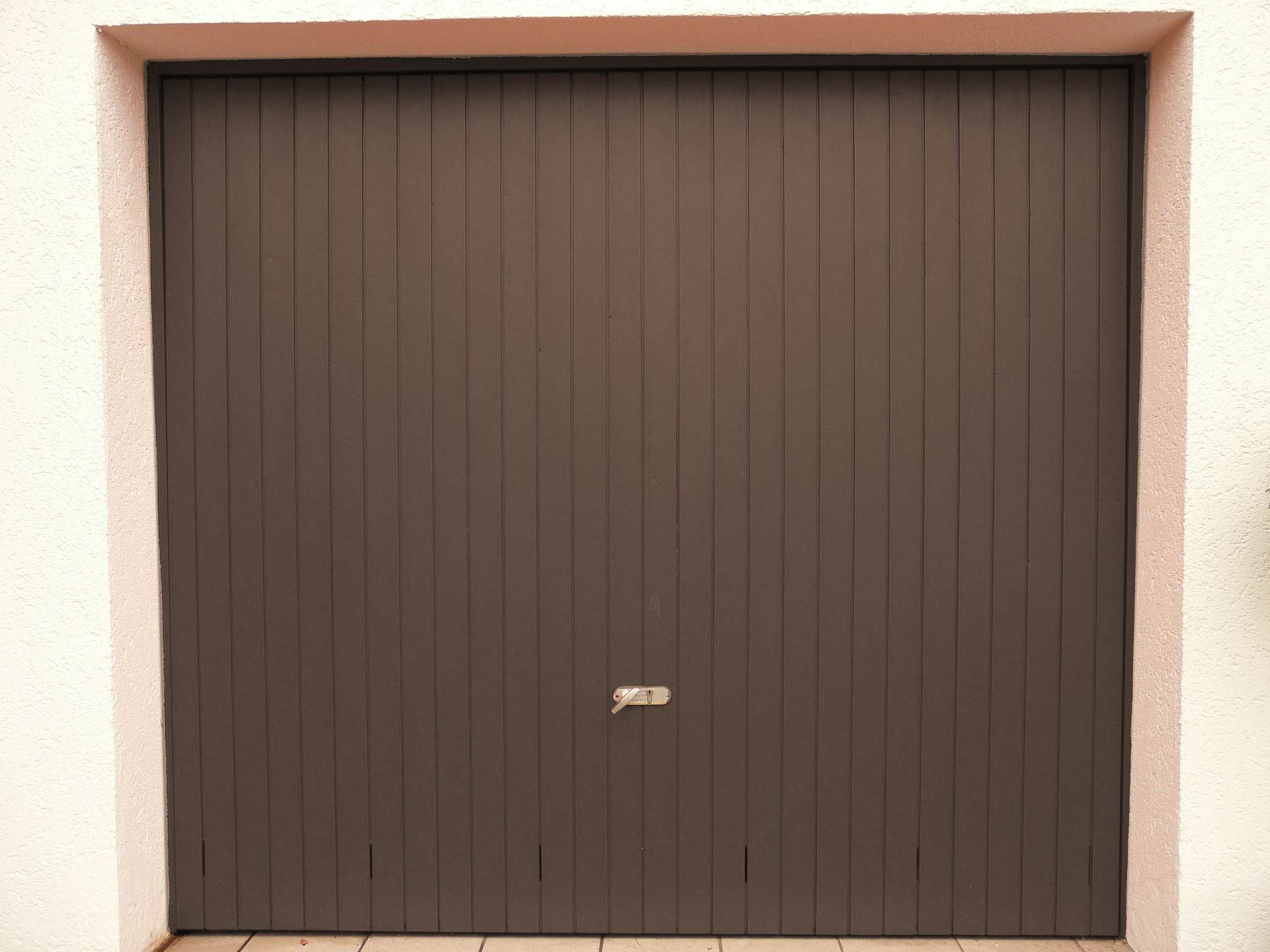 Porte de garage marron à rayures verticales, poignée centrale, encadrée d'un cadre beige clair.
