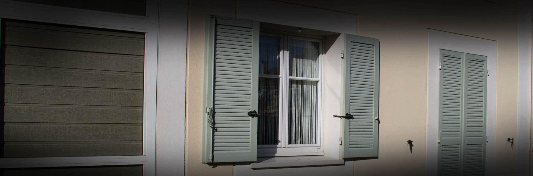 Vue extérieure d'un bâtiment avec trois fenêtres ; deux avec des volets, une ouverte avec des rideaux.
