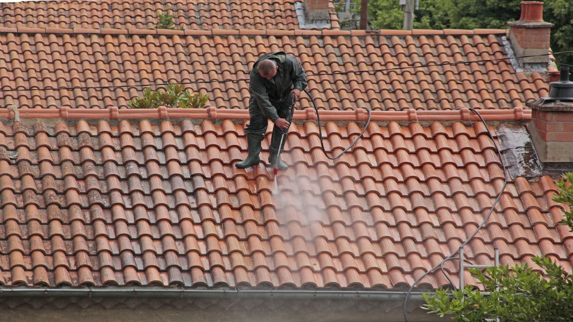 Homme vétu d'une combinaison de pluie, en train de nettoyer les tuiles d'une toiture avec un appareil haute pression