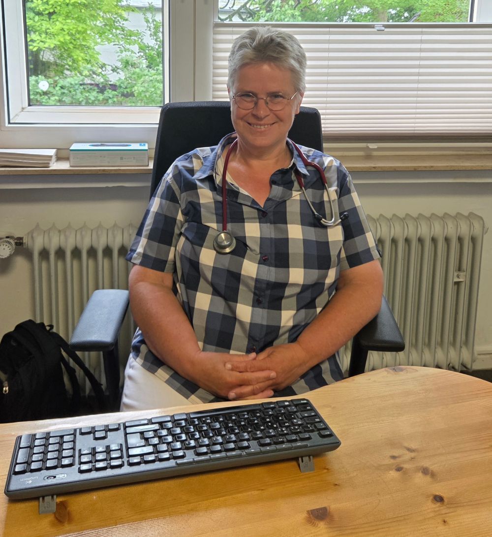 Eine lächelnde Person sitzt an einem Schreibtisch mit Tastatur, trägt ein kariertes Hemd und ein Stethoskop und befindet sich in einem Büro am Fenster.