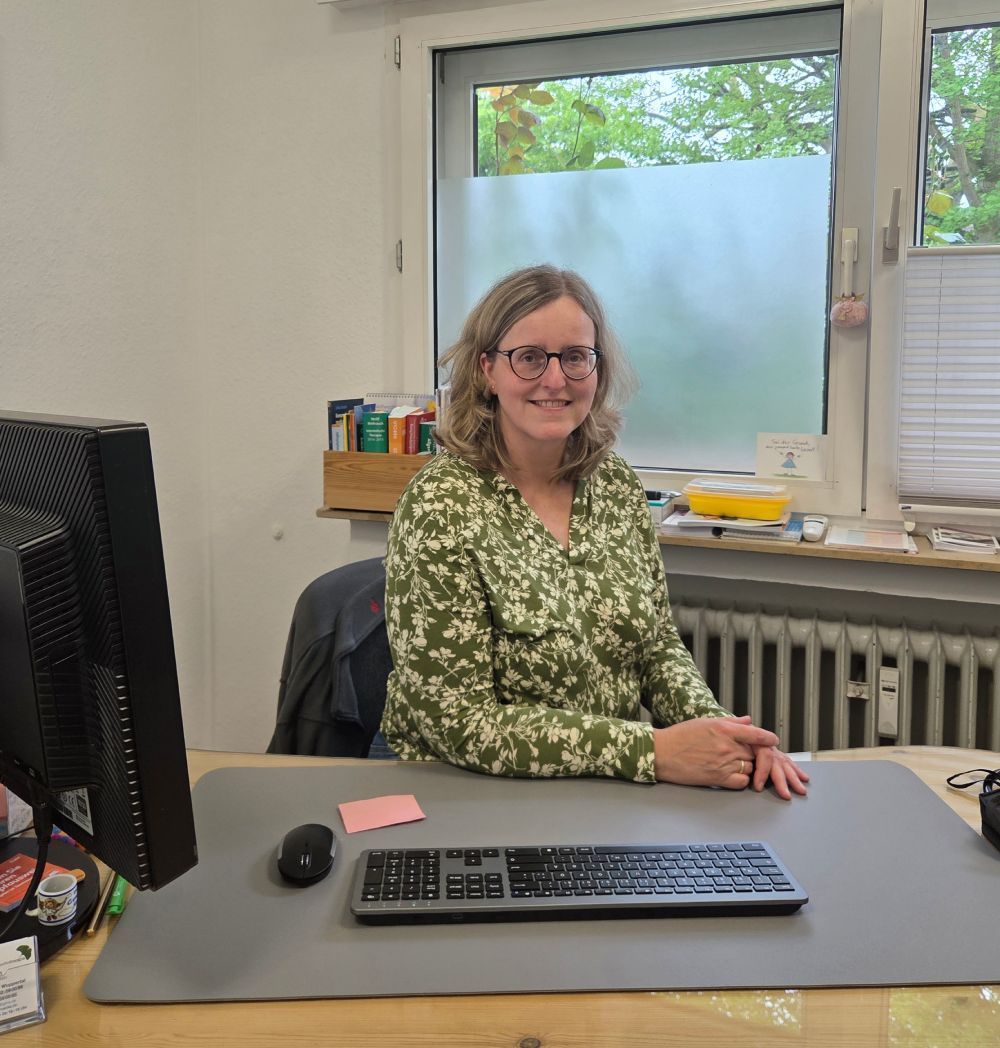 Frau lächelt an einem Schreibtisch in einem Büro; hinter ihr befinden sich ein Computerbildschirm, eine Tastatur und ein Fenster.