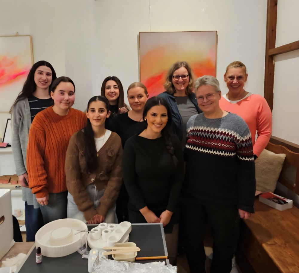 Eine Gruppe von Menschen posiert drinnen neben Schüsseln und Besteck; im Hintergrund hängt an der Wand ein farbenfrohes Kunstwerk.