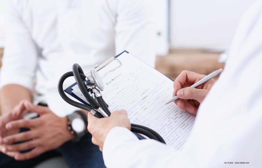 Der Arzt notiert auf einem Klemmbrett, während er einen sitzenden Patienten mit einer Blutdruckmanschette untersucht.