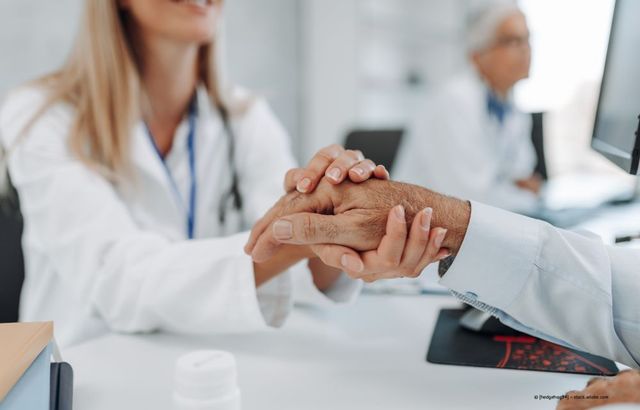Arzt und Patient schütteln sich in einem hellen Praxiszimmer die Hände; ein weiterer Arzt ist im Hintergrund verschwommen zu erkennen.
