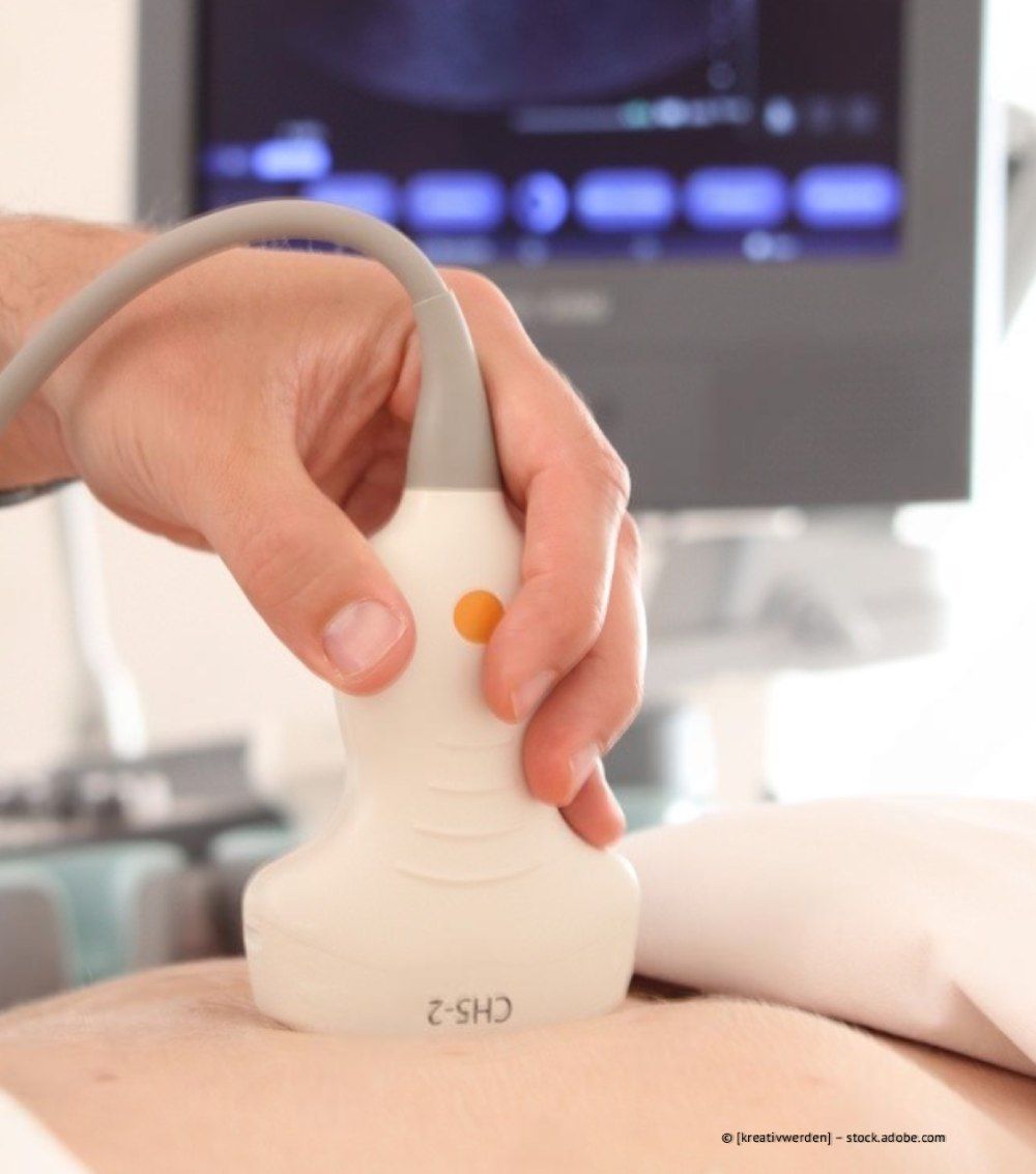 Eine Hand hält in einer klinischen Umgebung eine weiße Ultraschallsonde auf dem Bauch eines Patienten.
