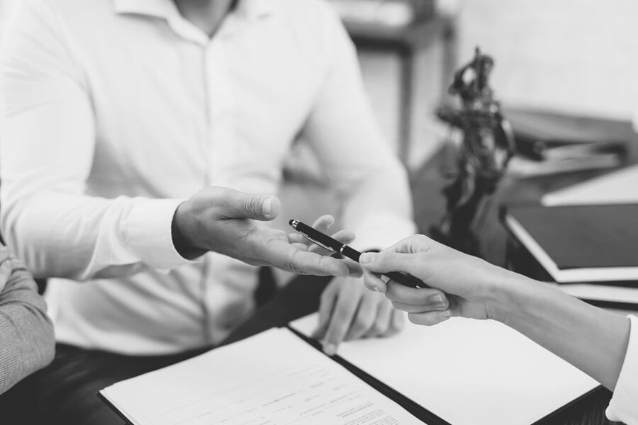 Pessoa entregando uma caneta para outra pessoa por meio de um documento. Ao fundo, uma estátua e um livro.
