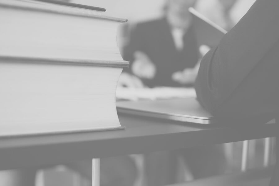 Pilha de livros sobre a mesa, pessoa com tablet em primeiro plano, duas pessoas desfocadas ao fundo, em um escritório.