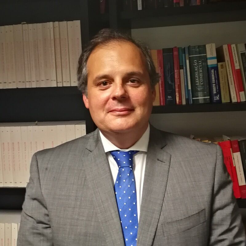 Homem de terno e gravata azul, sorrindo. Estantes de livros ao fundo.