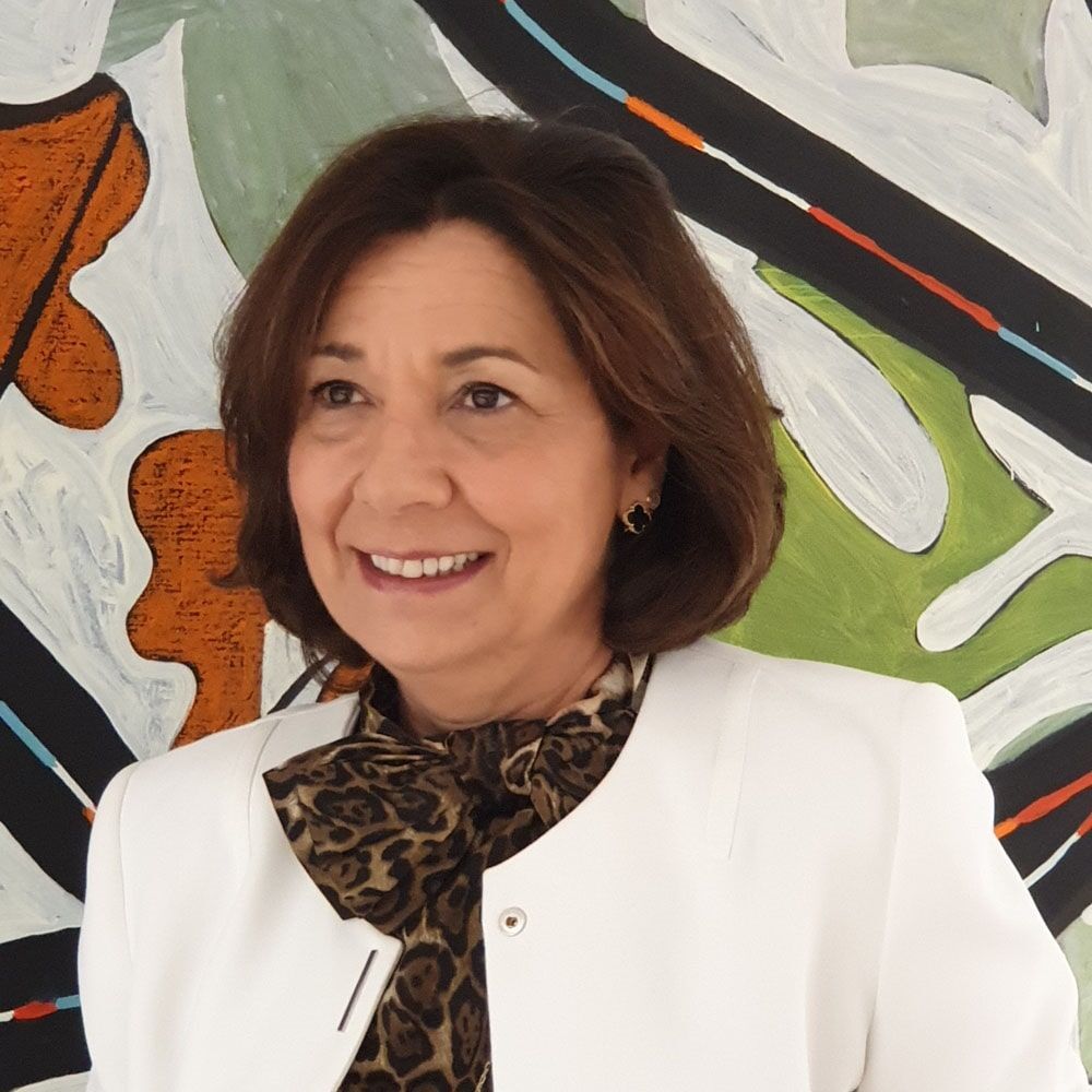 Mulher sorridente, vestindo um blazer branco e um lenço com estampa animal. Inclinada em frente a um fundo abstrato colorido.