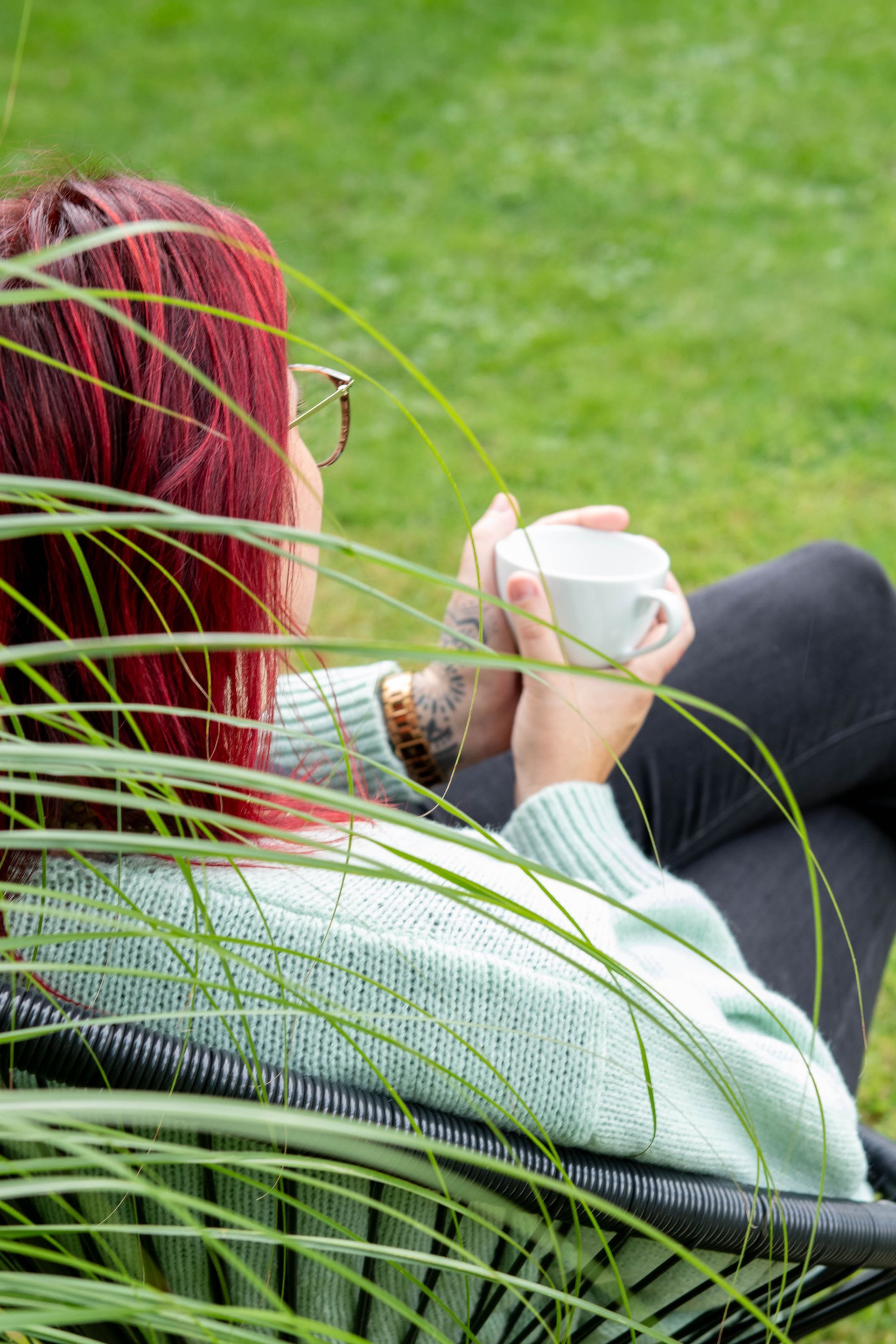 Eine Person mit Armtätowierungen formt mit ihren Händen ein Herz und blickt dabei auf eine grüne Landschaft.