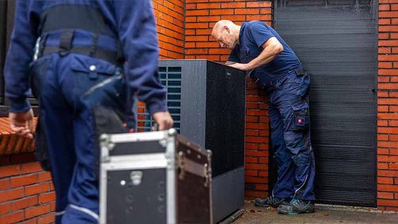 Zwei Männer arbeiten an einer Heizungsanlage außerhalb eines Backsteingebäudes.