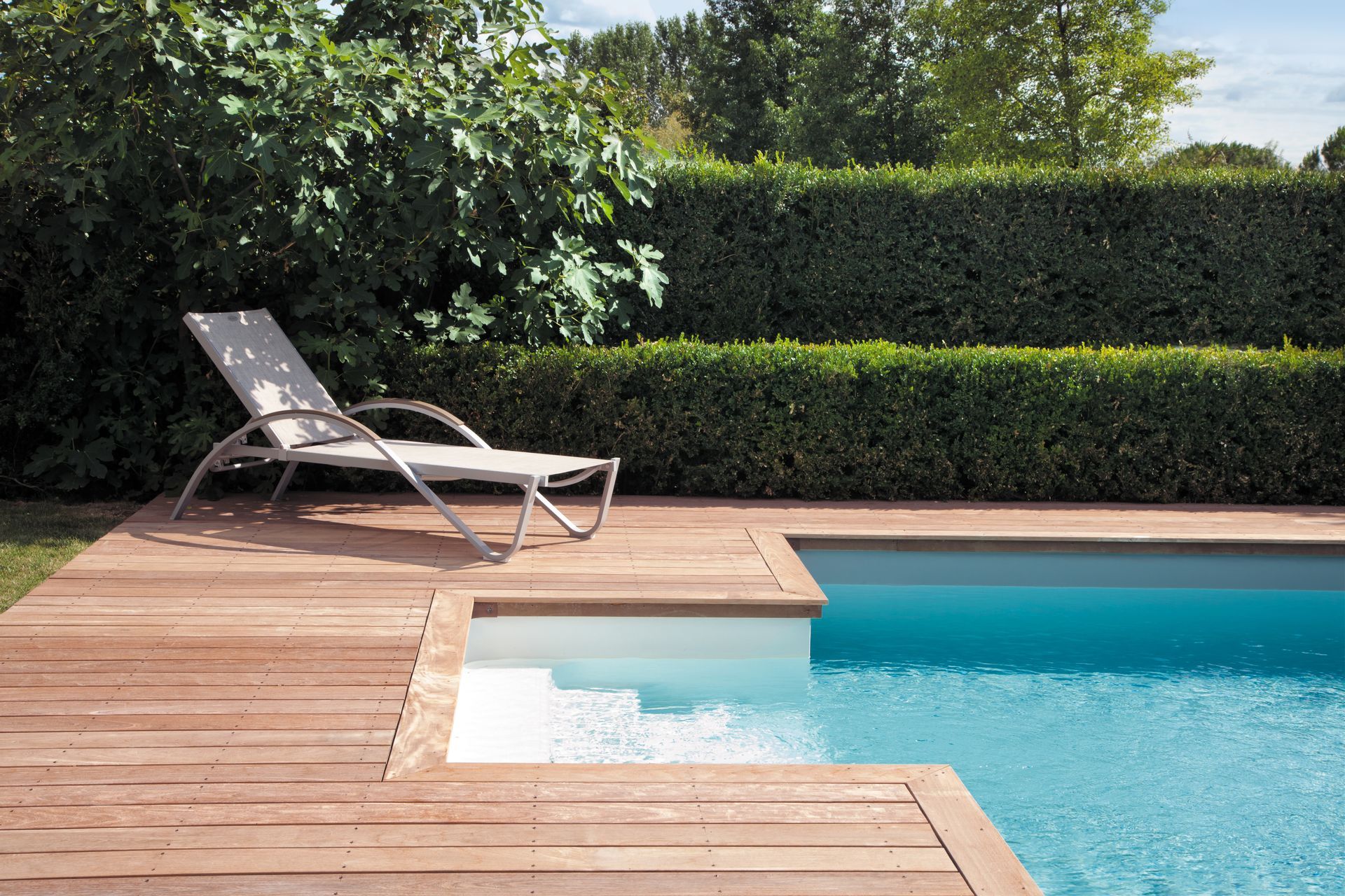 Une piscine avec terrasse en bois et chaise longue, bordée d'une haie.