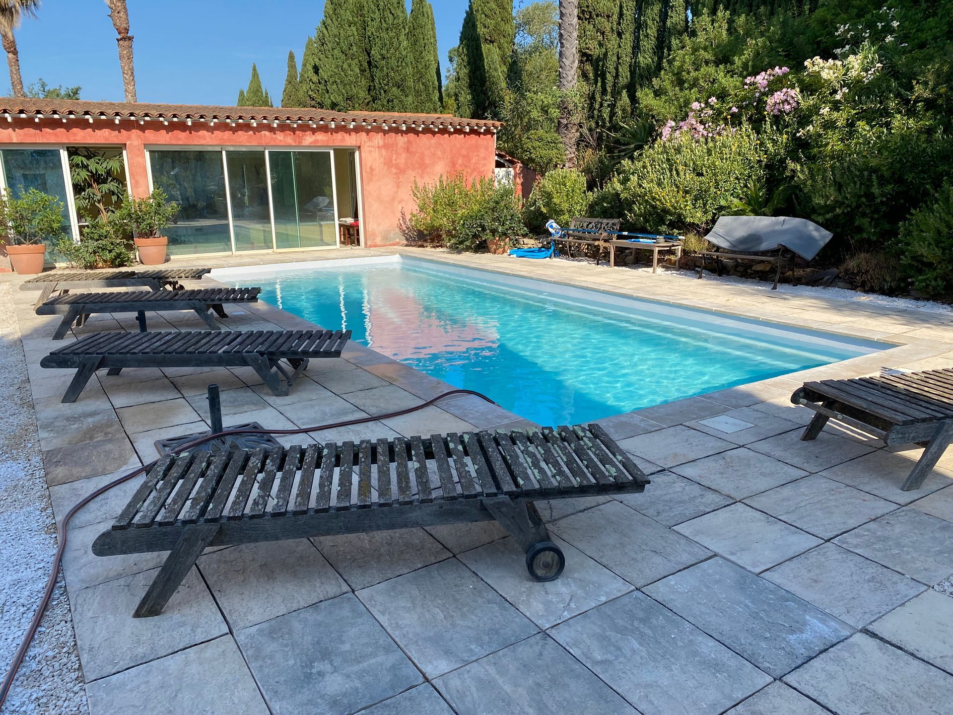 Piscine avec chaises longues et maison rouge dans un cadre ensoleillé.