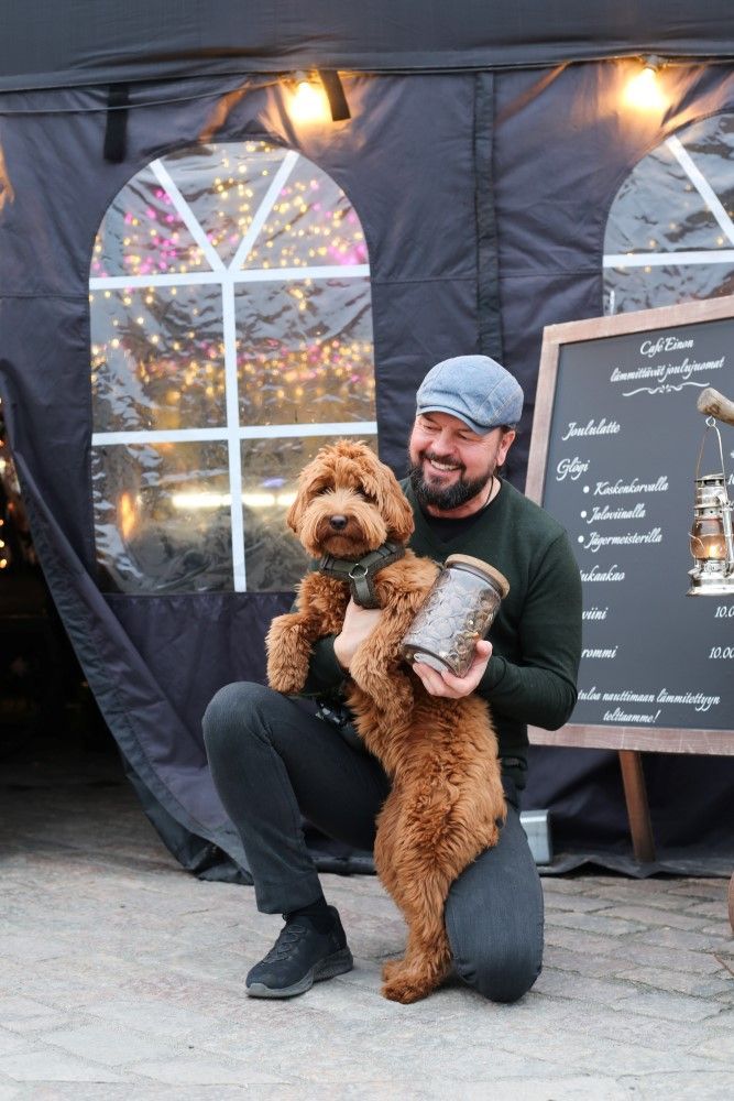 Omistaja polvilla, kädessä Rovio koirankeksipurkki ja sylissä pientä ruskeaa koira, kahvilan oma koira Pasanen.
