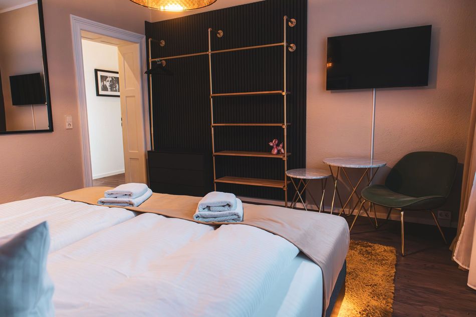 Blick auf ein Bett mit Handtüchern, ein offenes goldenes Regal, einen grünen Sessel und einen Wandfernseher.
