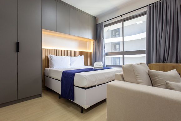 Quarto moderno com cama arrumada, guarda-roupa embutido, sofá e janela grande com cortinas cinza.