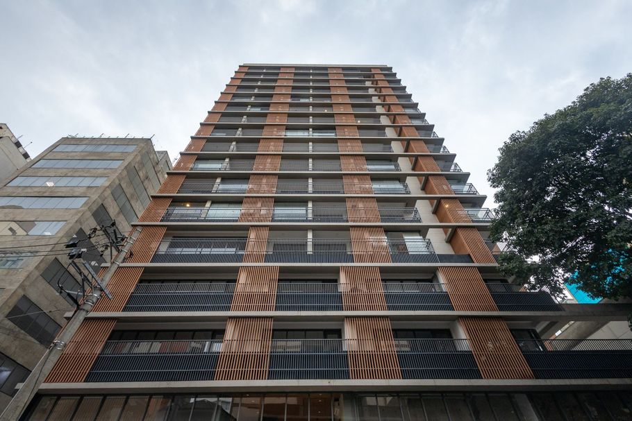 Vista em ângulo baixo de um prédio de apartamentos moderno e alto, com detalhes em laranja e varandas, contra um céu cinzento.