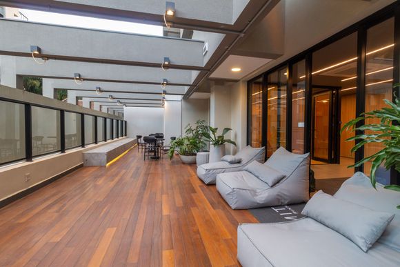 Lounge moderno na cobertura com deck de madeira, pufes cinza e área de estar com paredes de vidro.