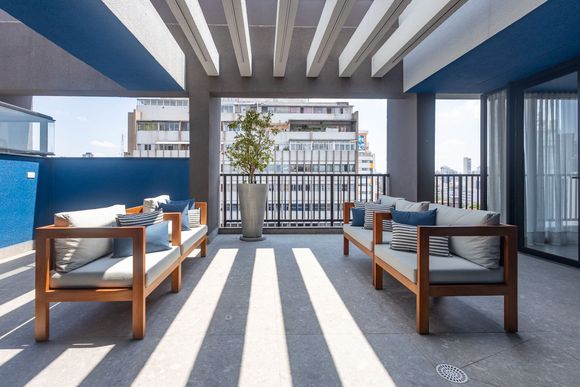 Lounge moderno na cobertura com sofás de madeira, almofadas azuis e vista para o horizonte da cidade sob sombras listradas.