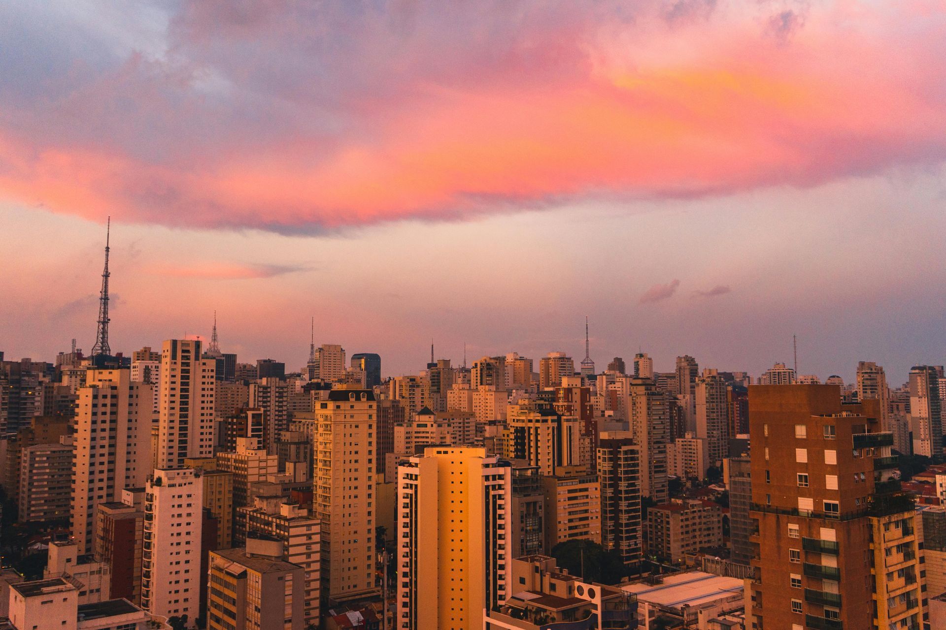 Vista da cidade ao pôr do sol com nuvens rosa-alaranjadas sobre densos arranha-céus.