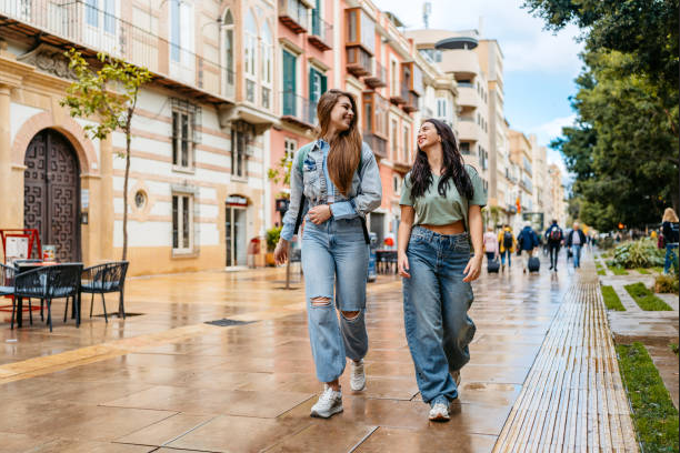Dos mujeres con vaqueros caminan por una calle mojada de la ciudad, bordeada de edificios y árboles.