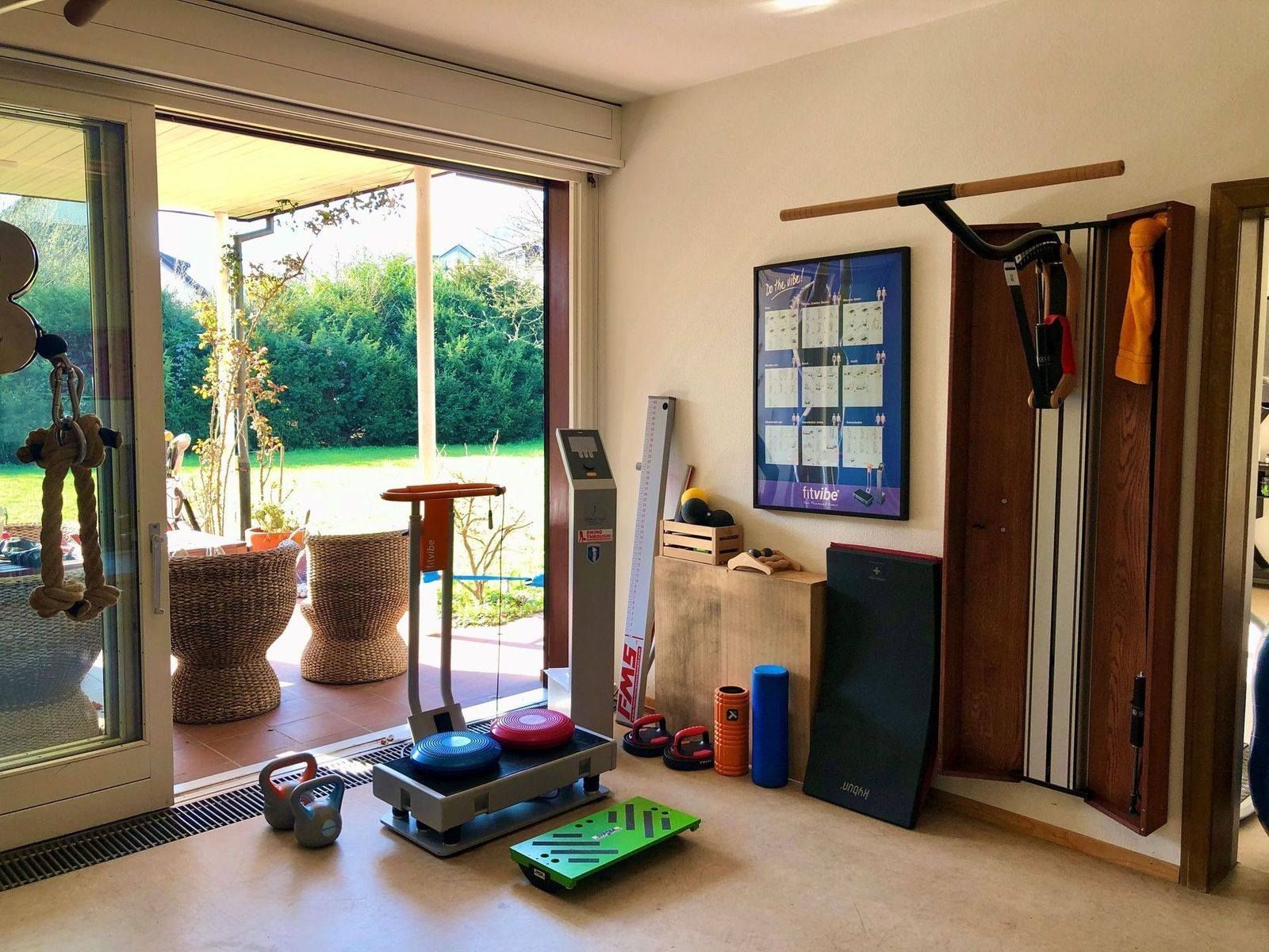 Kleiner Fitnessraum mit Trainingsgeräten, Gewichten, Sprossenwänden und Blick auf einen sonnigen Garten durch Glastüren.