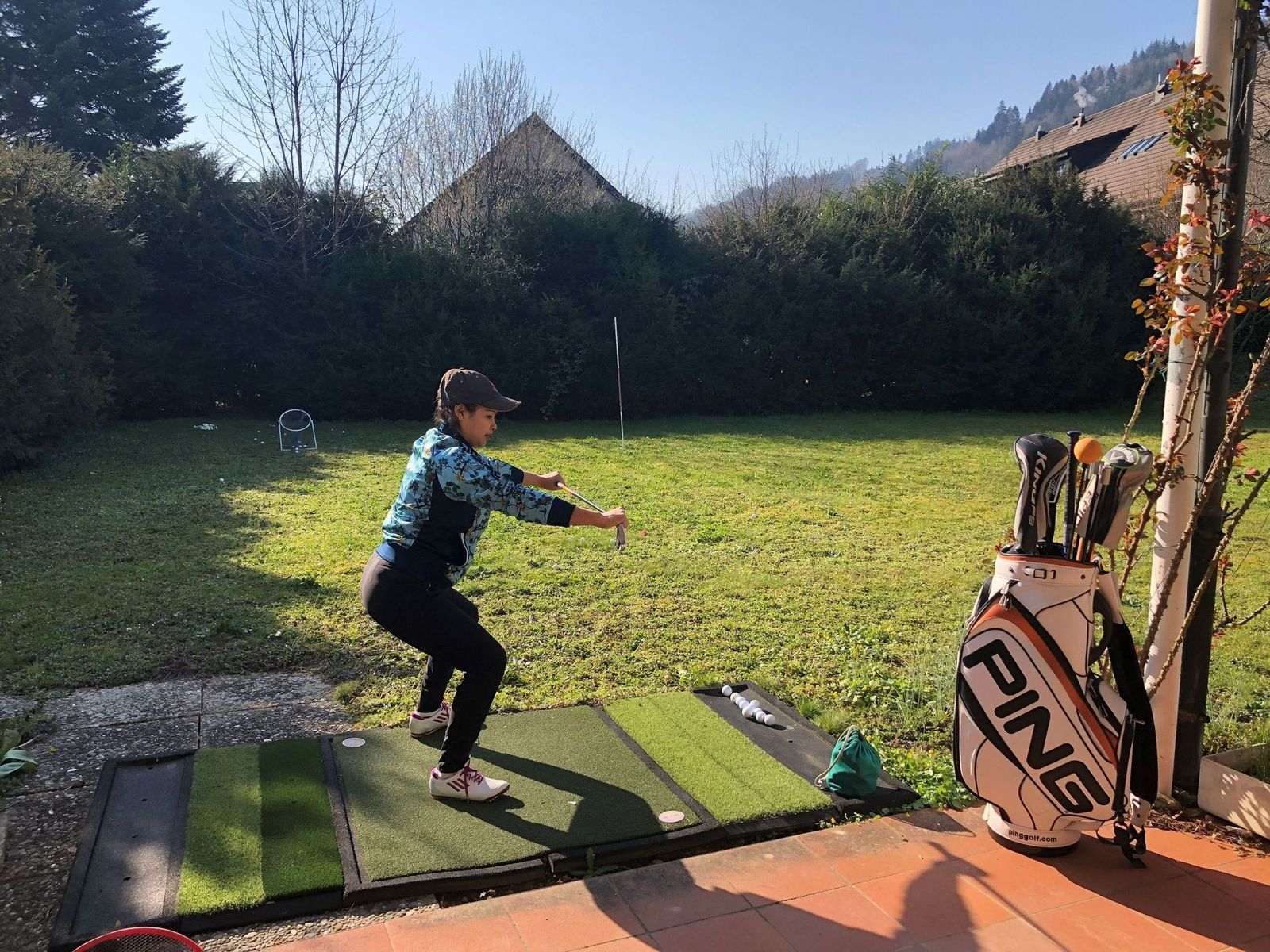 Eine Person übt in einem sonnigen Garten auf einer Matte ihren Golfschwung; eine Golftasche steht in der Nähe.