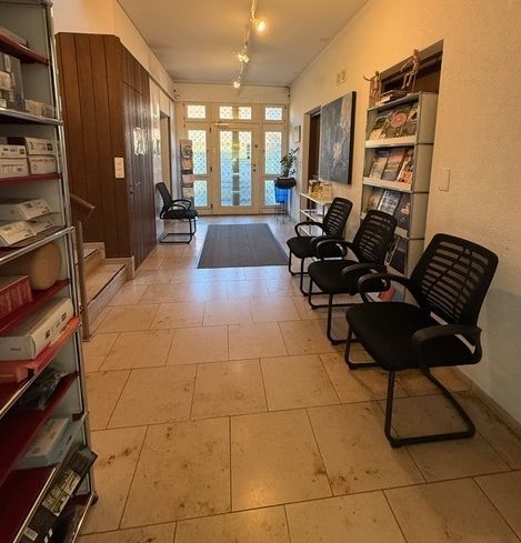 Schmaler Warteraum einer Klinik mit schwarzen Stühlen, Wandregalen und Glastüren am Ende.