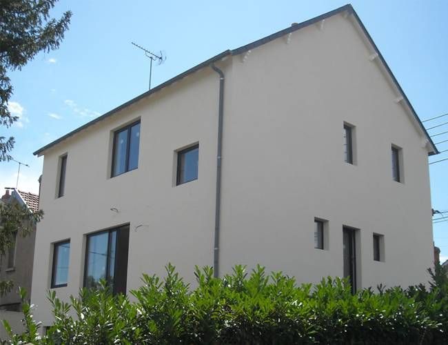 Maison moderne beige à deux étages avec des fenêtres à encadrement bleu derrière des arbustes verts