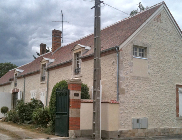 Maisonnette en pierre beige avec toit de tuiles rouges, cheminée en briques et portail vert à côté d'un poteau électrique.