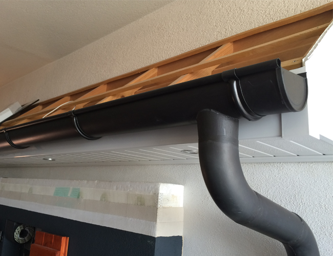 Gouttière et descente pluviale noires sous un avant-toit en bois, contre un mur extérieur clair.