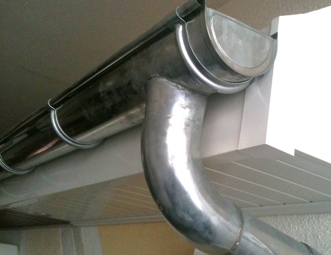 Tuyaux de gouttière métalliques reliés à l'avant-toit d'une maison, avec une descente de gouttière courbée et une ligne de toit visible