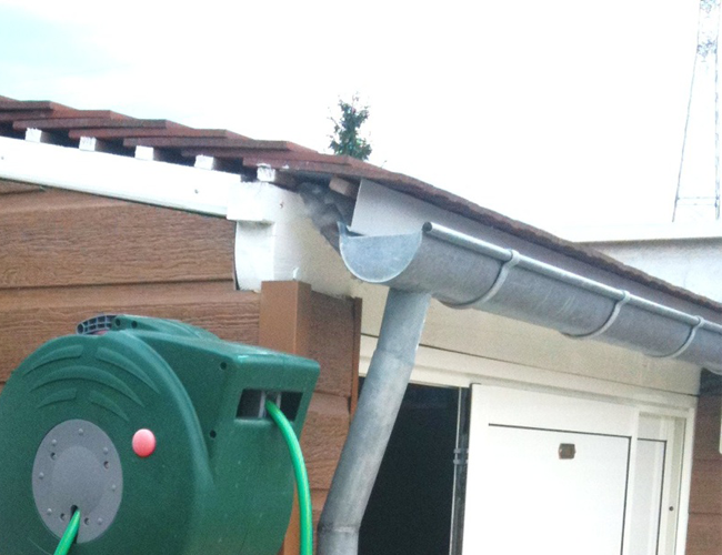 Un enrouleur de tuyau d'arrosage vert se trouve à côté d'un bâtiment blanc avec une gouttière et une descente pluviale grises sous un toit de tuiles.