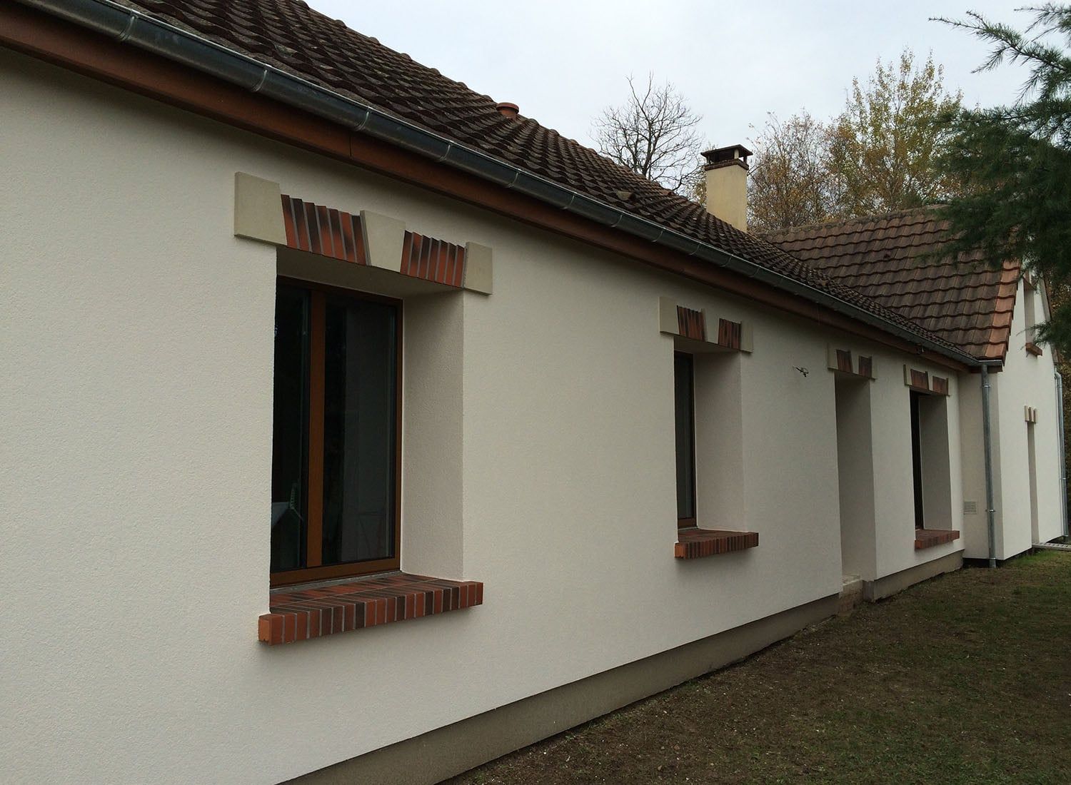 Maison blanche avec fenêtres encadrées de briques et toit de tuiles donnant sur une cour gazonnée