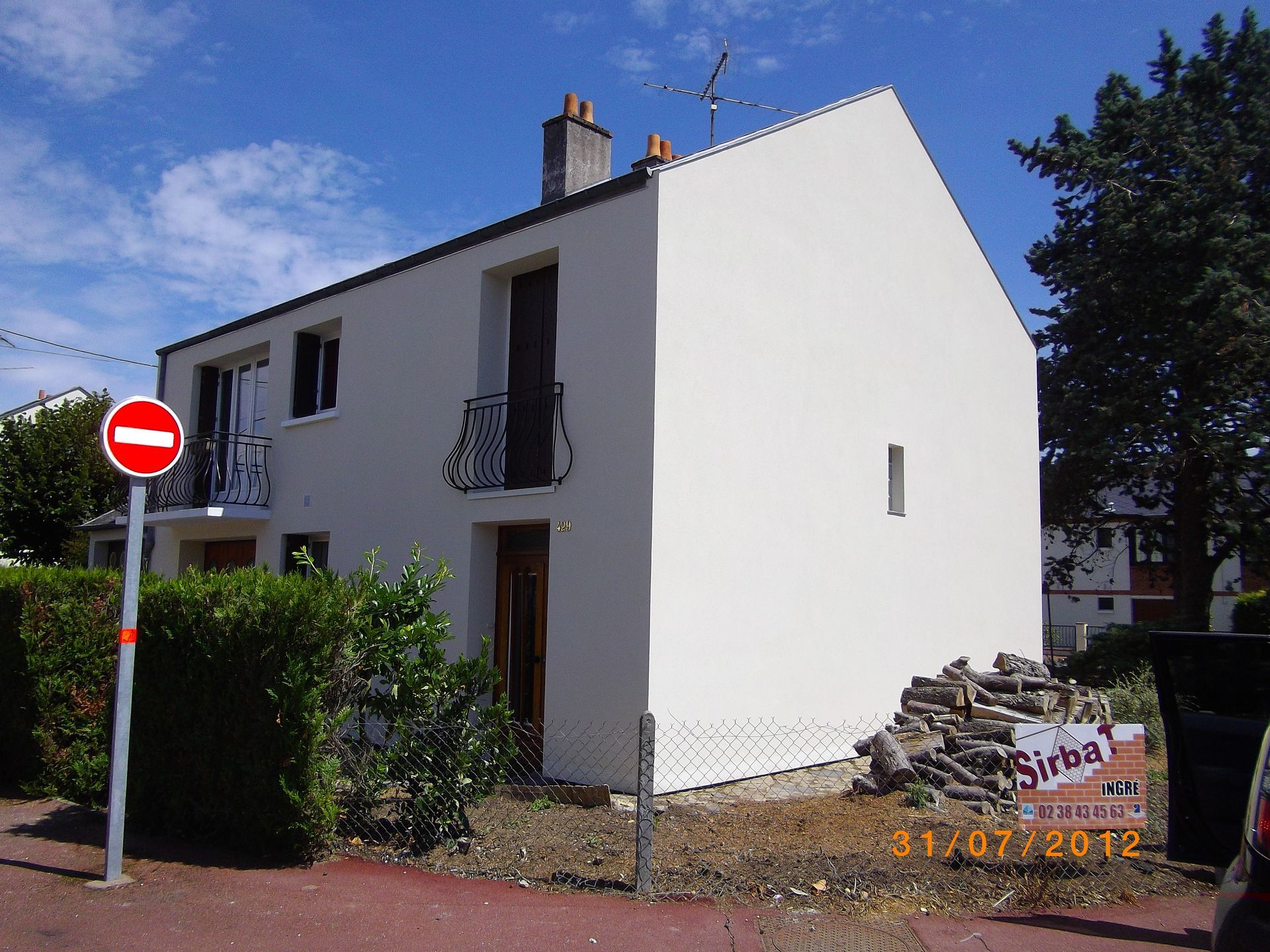 Maison blanche à deux étages avec un petit balcon, à côté d'un panneau « Entrée interdite » et d'une haie sous un ciel bleu.