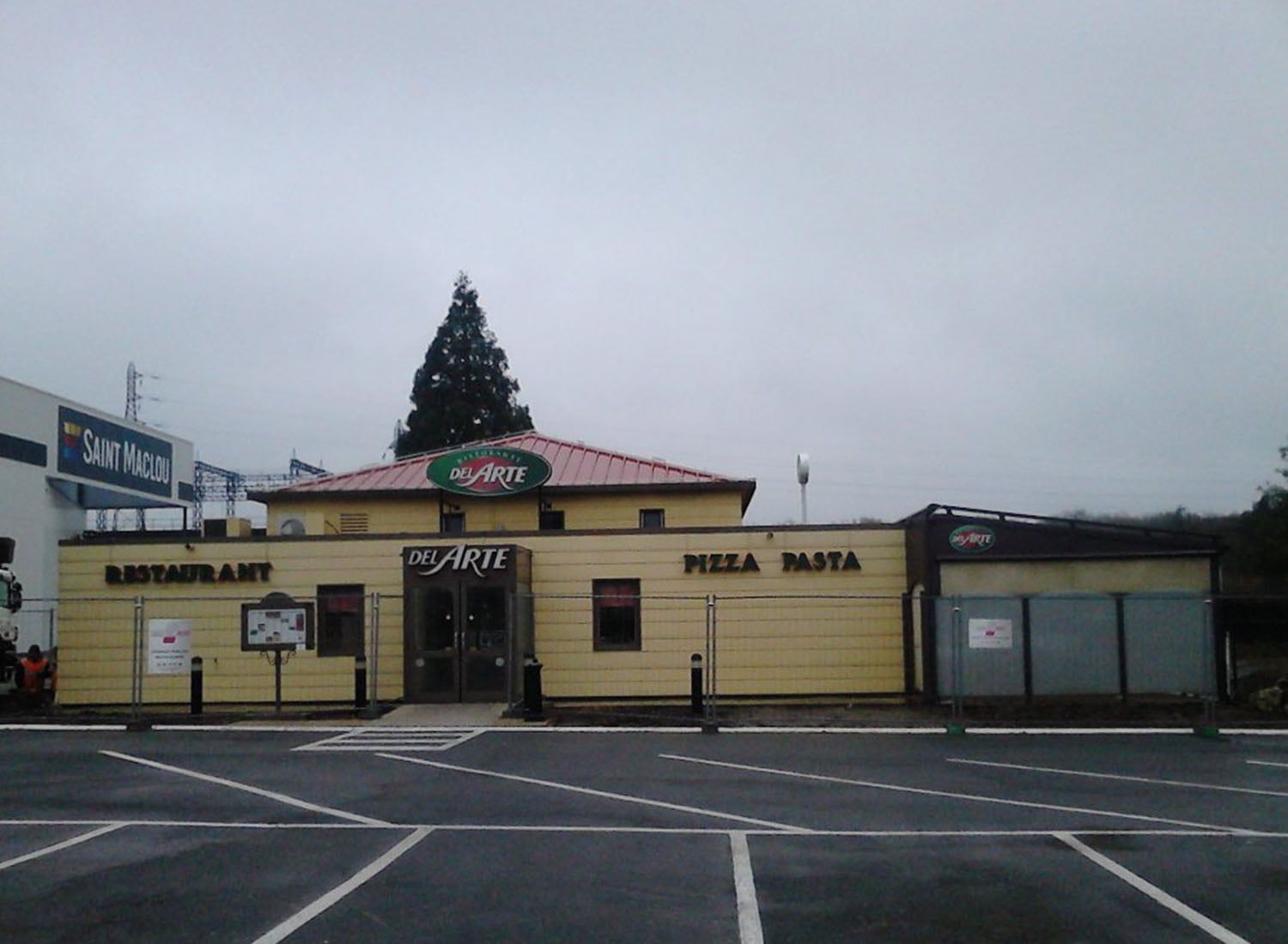 Petit bâtiment jaune abritant un restaurant, avec des enseignes, une entrée couverte et un parking vide sous un ciel nuageux.