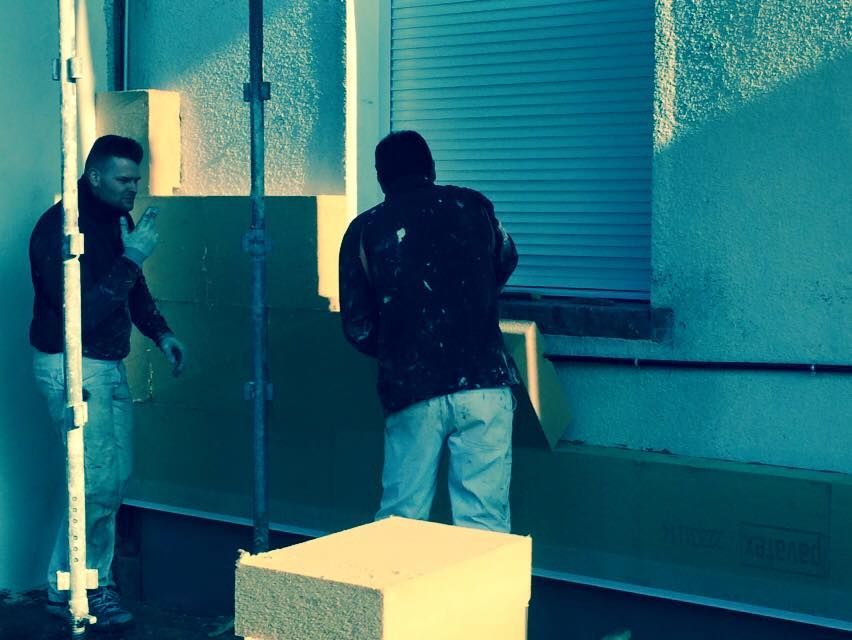 Deux personnes déplacent des cartons dans une pièce ressemblant à un entrepôt, avec des murs turquoise et une fenêtre aux volets fermés.